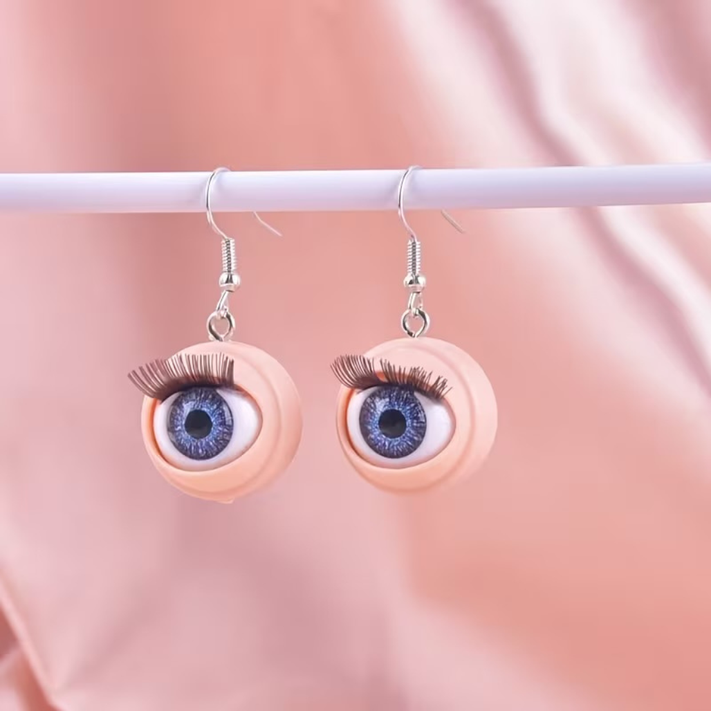 Eyeball-shaped earrings hang on a simple earring display against a pink background