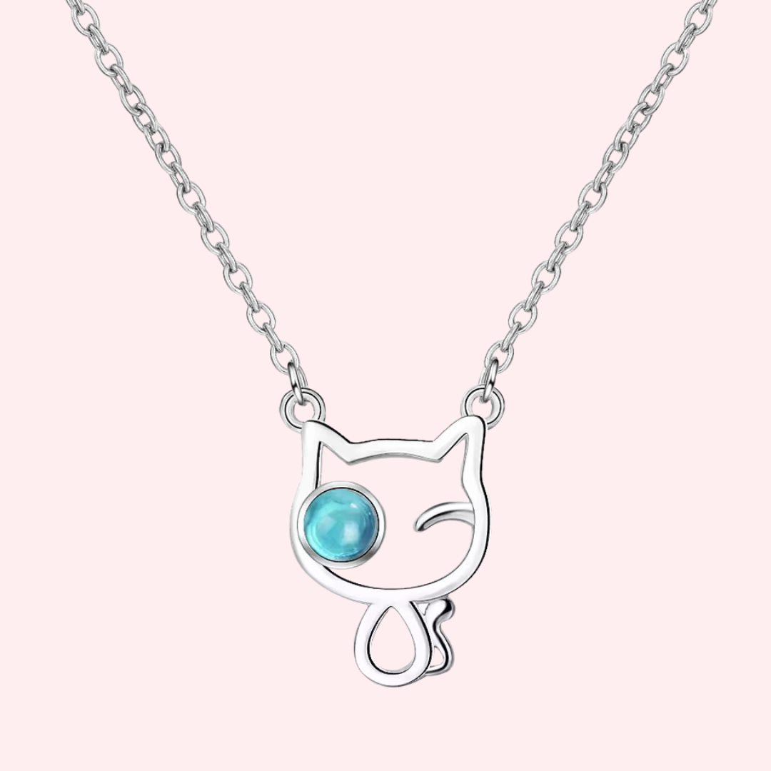 Silver cat dangles on a delicate chain. One cat eye is round and blue and the other is winking. Curled cat tail.