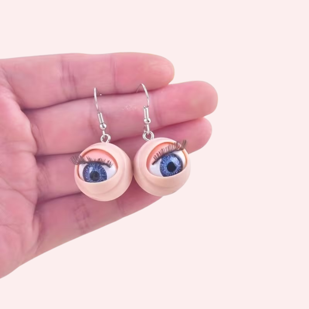 Eyeball-shaped earrings held in a hand against a light pink background