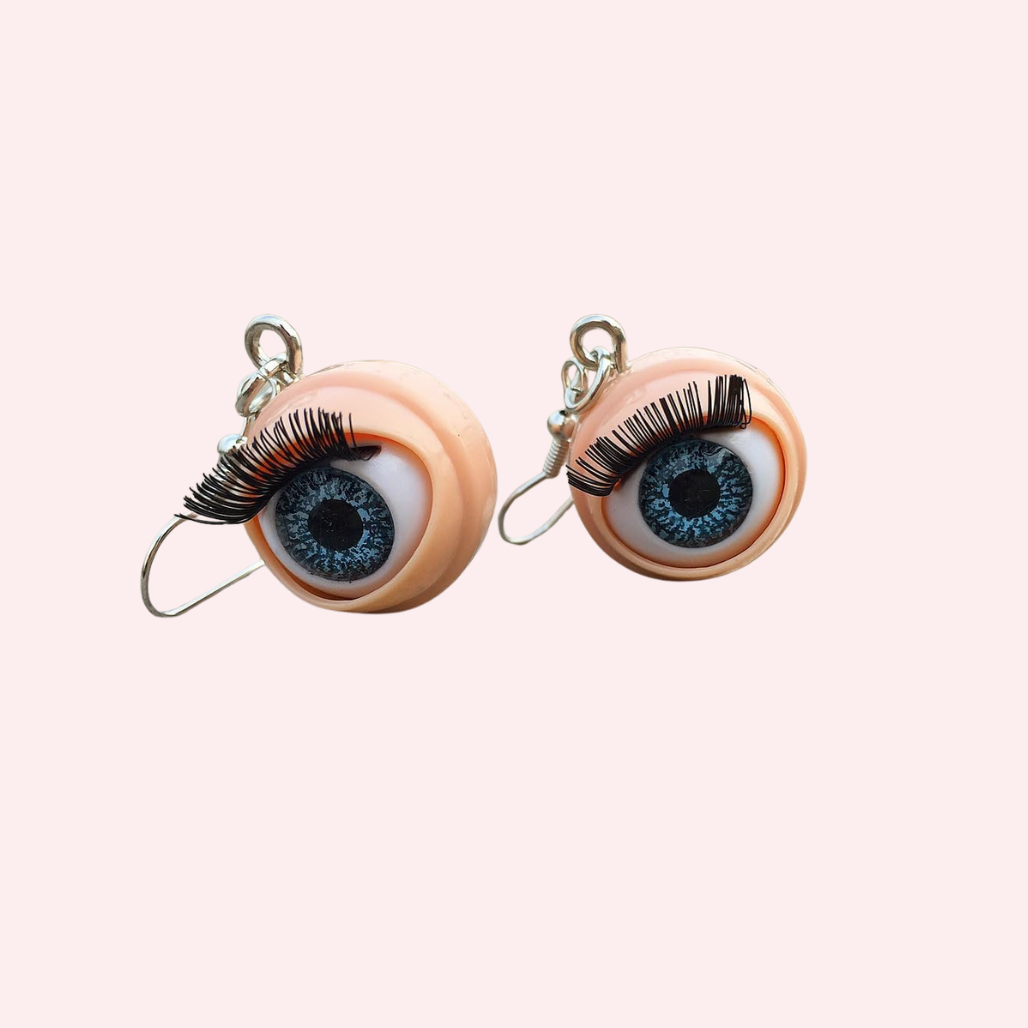 Round eyeball earrings with false lashes and blue eyes against a light pink background