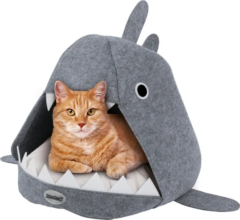 Grey felt cat bed shaped like a shark. An orange cat peeks out of the shark’s mouth with small white felt teeth looking like it might eat the cat. 