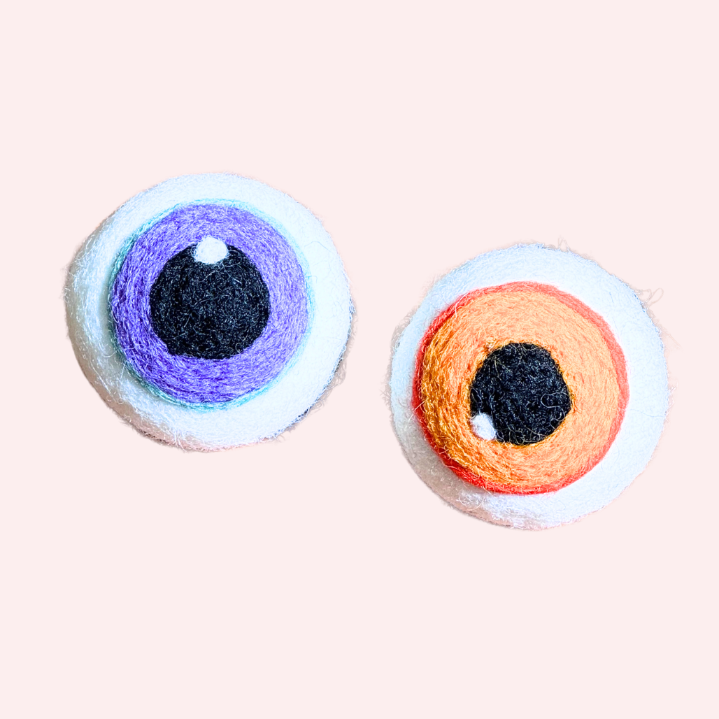 Two handmade felt wool eyes with different colored irises, one purple and one orange, displayed on a pale pink background.